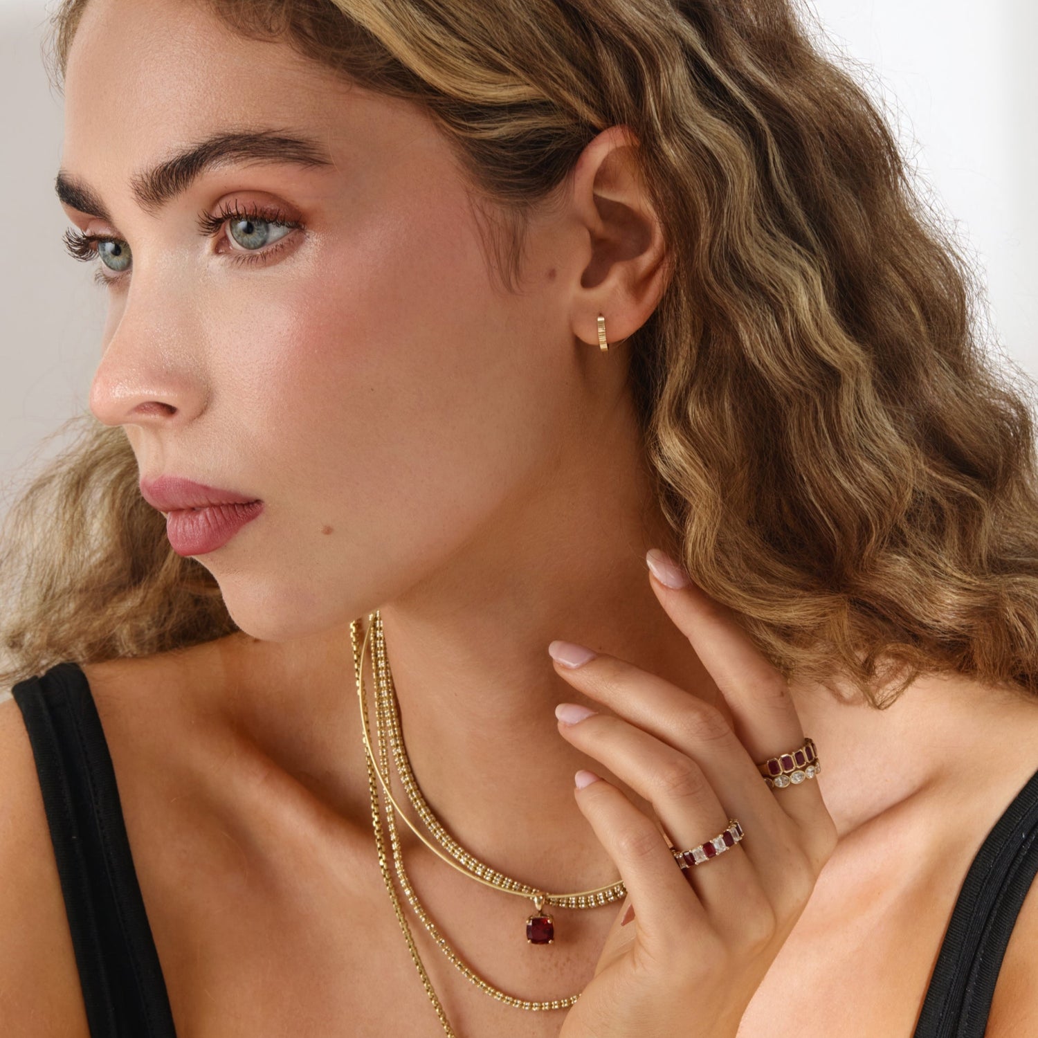 Woman wearing gold jewelry including necklaces, earrings, and rings on a plain background