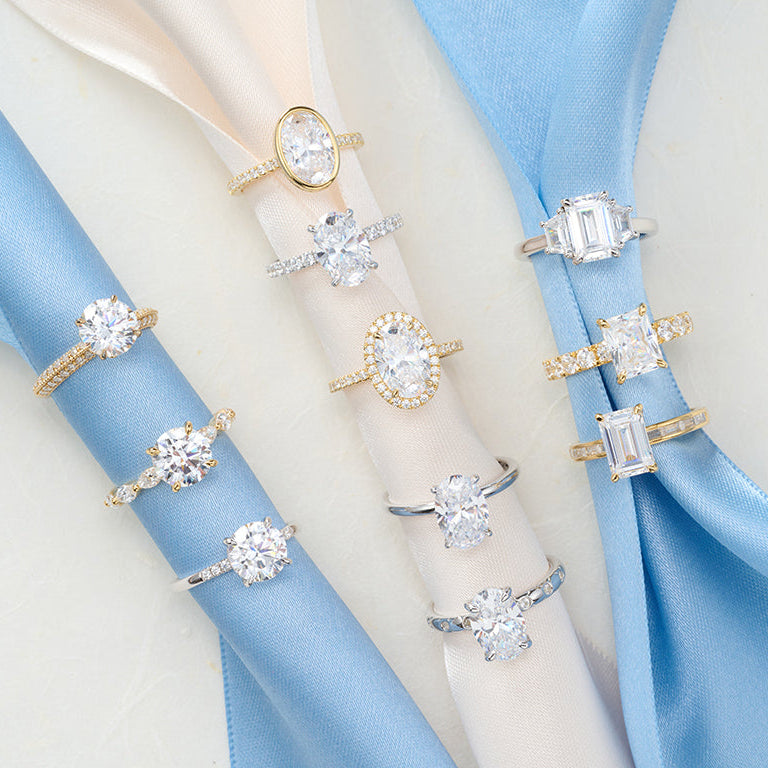 Lab-grown diamond engagement rings in round, oval, emerald and radiant cuts displayed on blue and ivory ribbon.